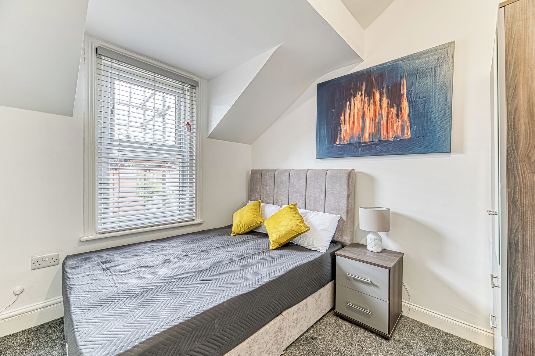 A bedroom with a bed, bedside table, lamp, and a painting of a fire.