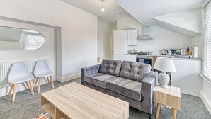 Un salon moderne avec un canapé gris, deux fauteuils blancs, une table basse en bois et un coin cuisine équipé d’appareils électroménagers et doté d’une fenêtre avec des stores.