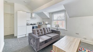 Un appartement moderne avec un canapé gris, une table basse en bois, une cuisine avec des meubles blancs et une grande fenêtre équipée de stores.