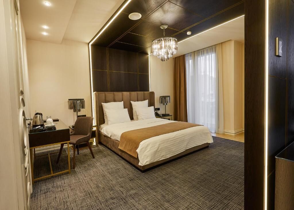 A modern hotel room with a large bed, a desk with a chair, a chandelier, and a view of the outdoors.