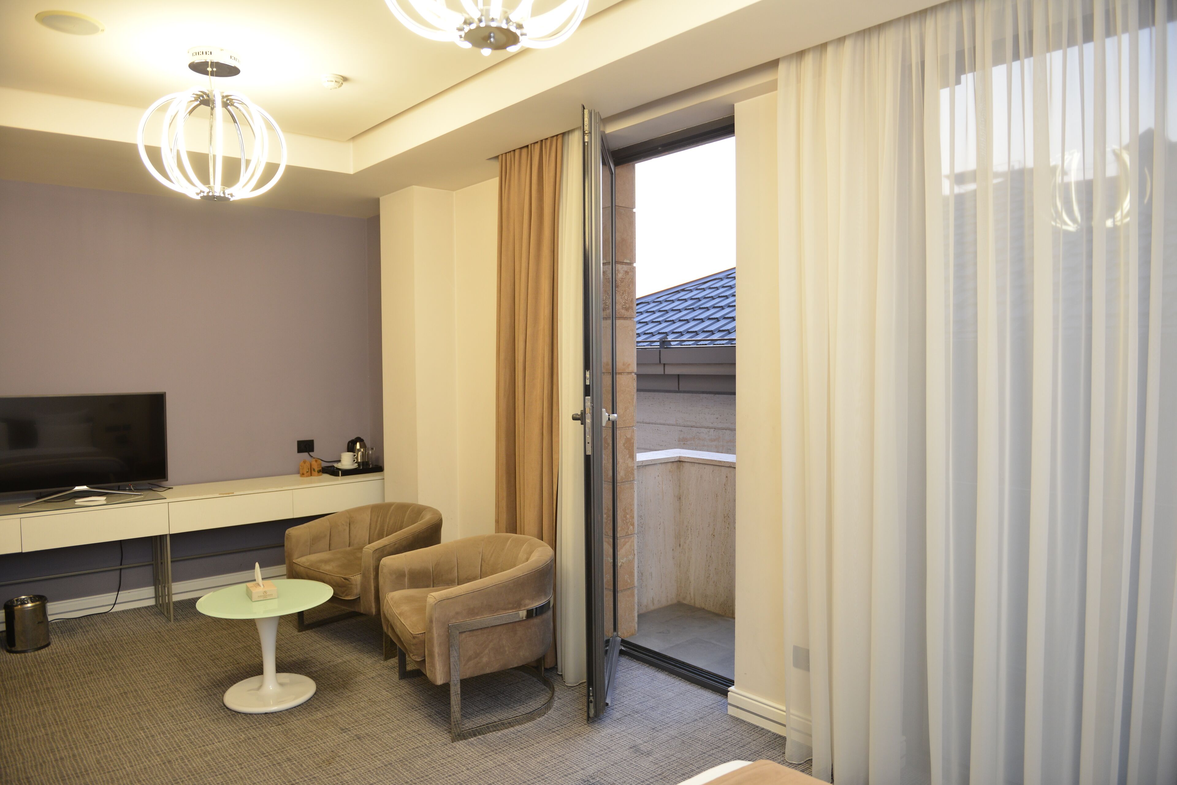 A modern hotel room with a flat-screen TV, a small round table, two armchairs, and a sliding glass door leading to a balcony.
