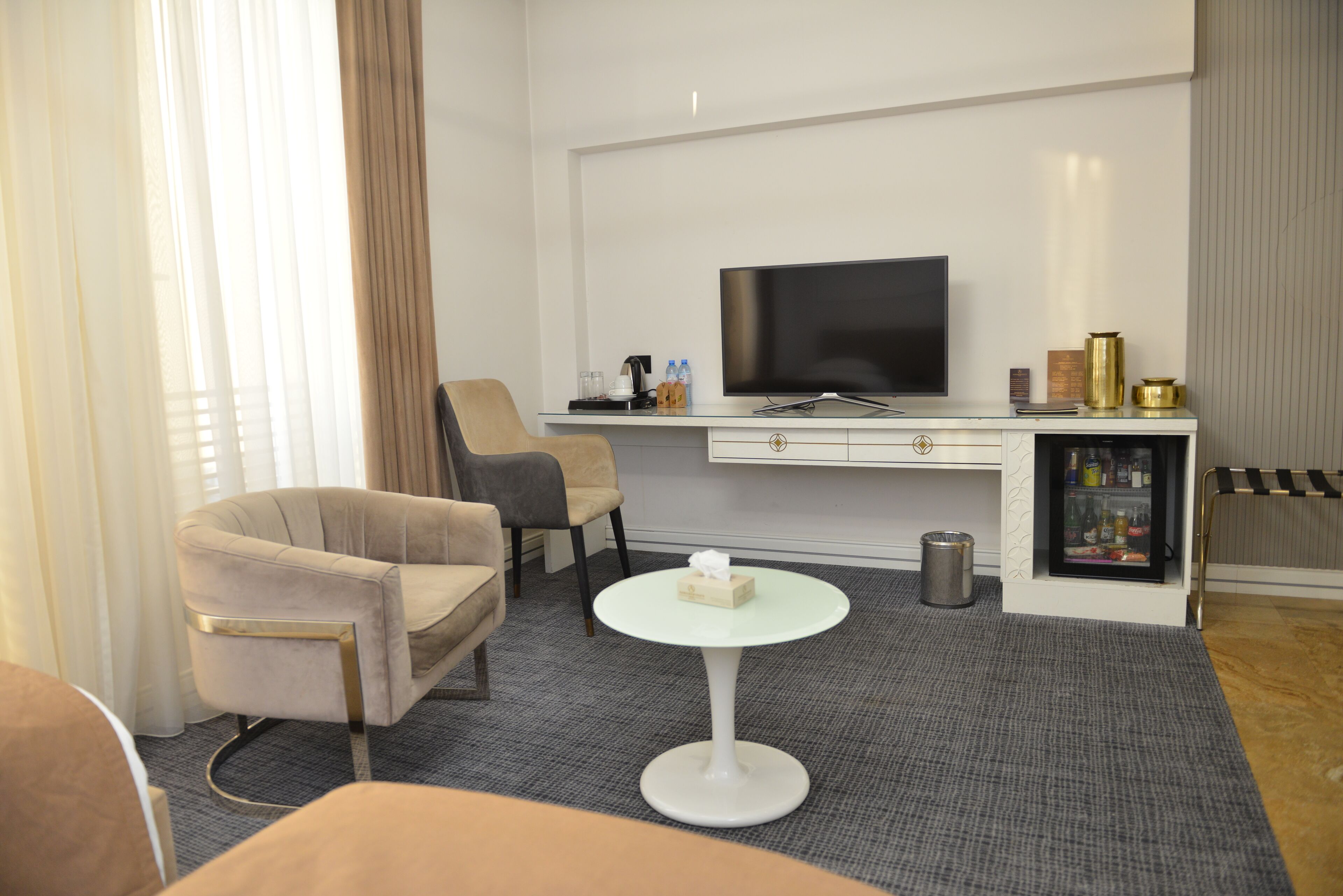 A modern hotel room with a flat-screen TV, a sofa, a coffee table, a minibar, and a large window with curtains.
