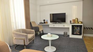A modern hotel room with a flat-screen TV, a sofa, a coffee table, a minibar, and a large window with curtains.