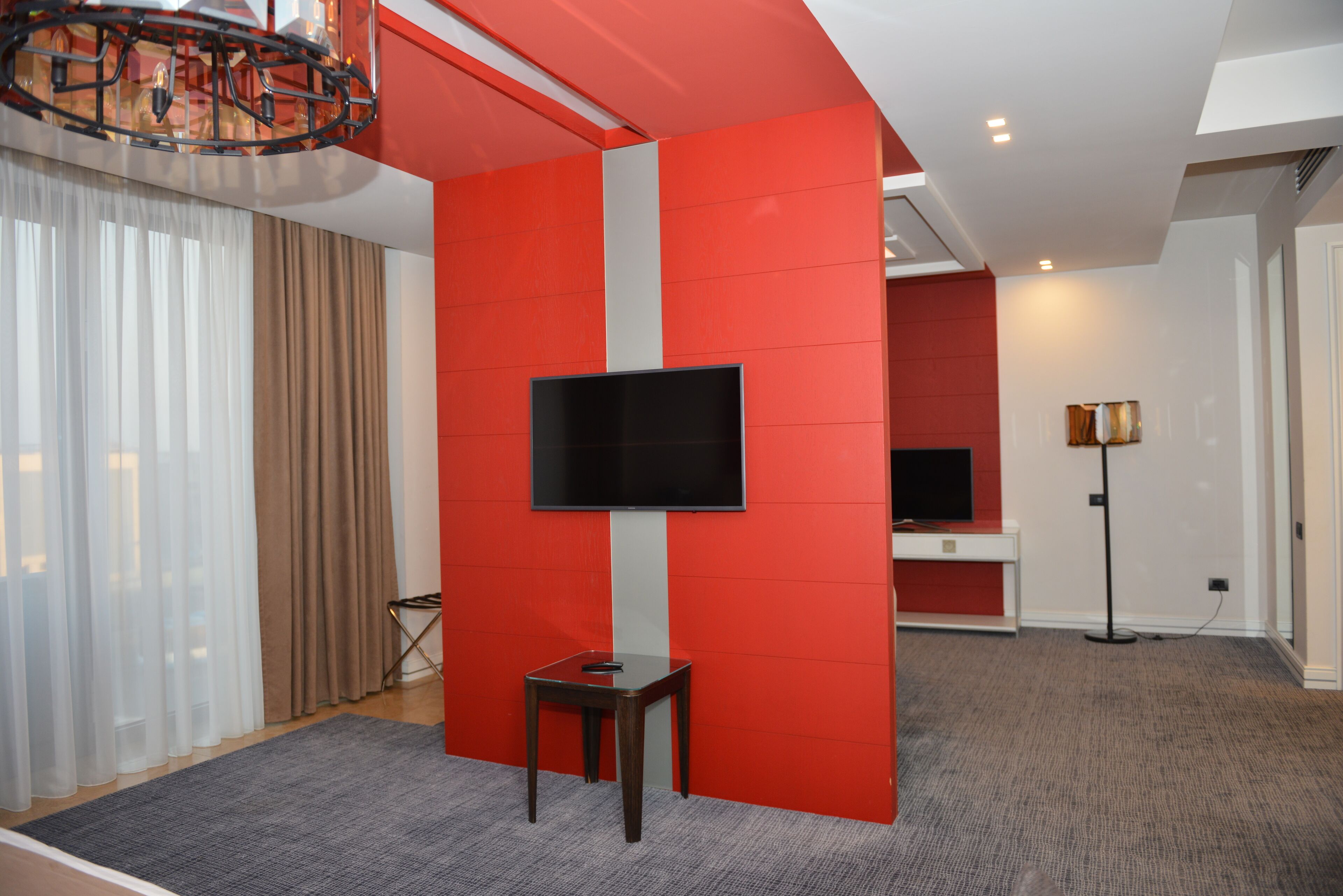 A modern hotel room with a large flat-screen TV mounted on a red accent wall, a small wooden table, and a grey carpet.