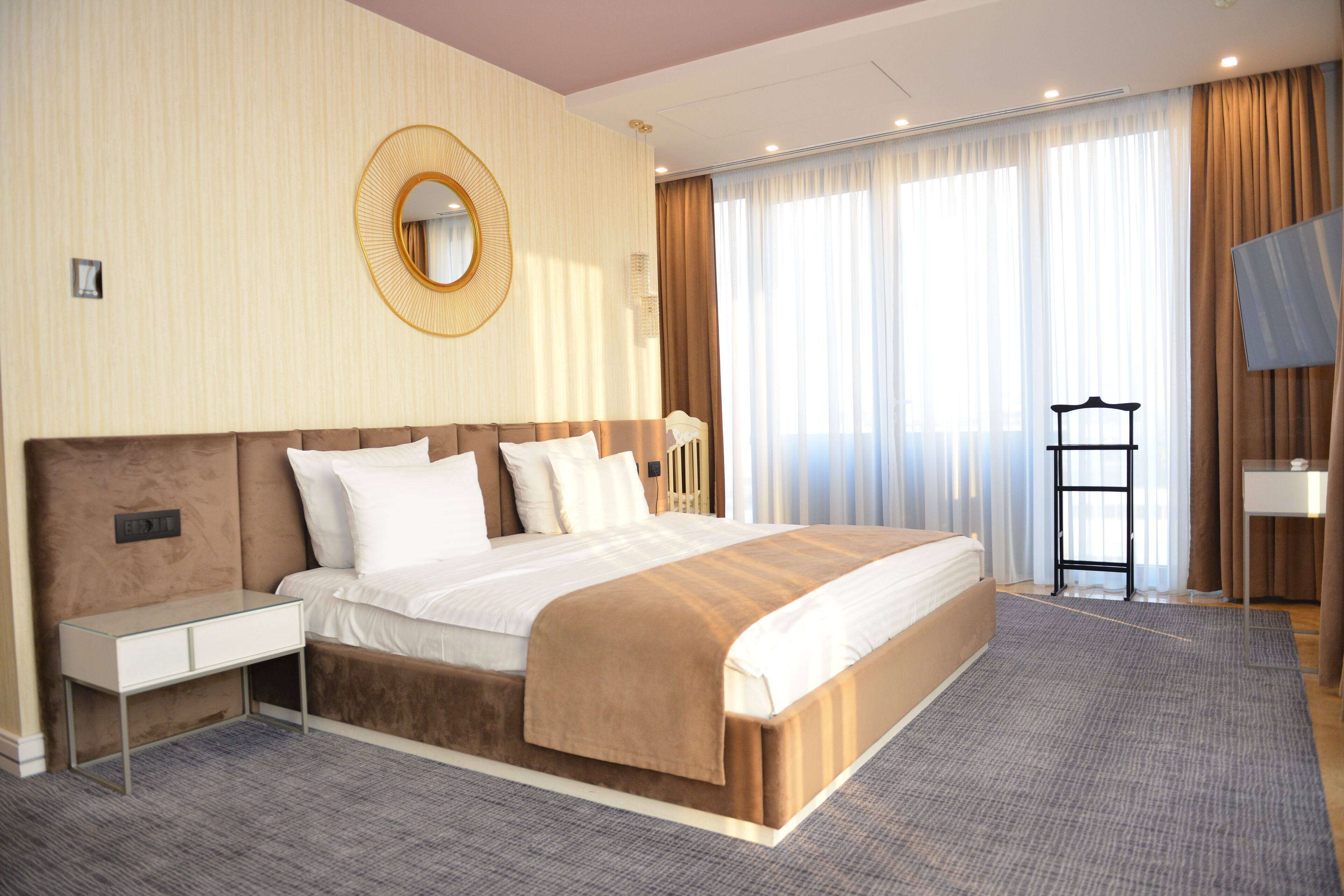 Habitación de hotel con una cama grande, una mesita de noche, un espejo, un televisor y una ventana con cortinas.