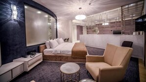 A modern hotel room with a curved ceiling, a bed with a headboard, a seating area with a chair and a small round table, and a kitchenette in the background.