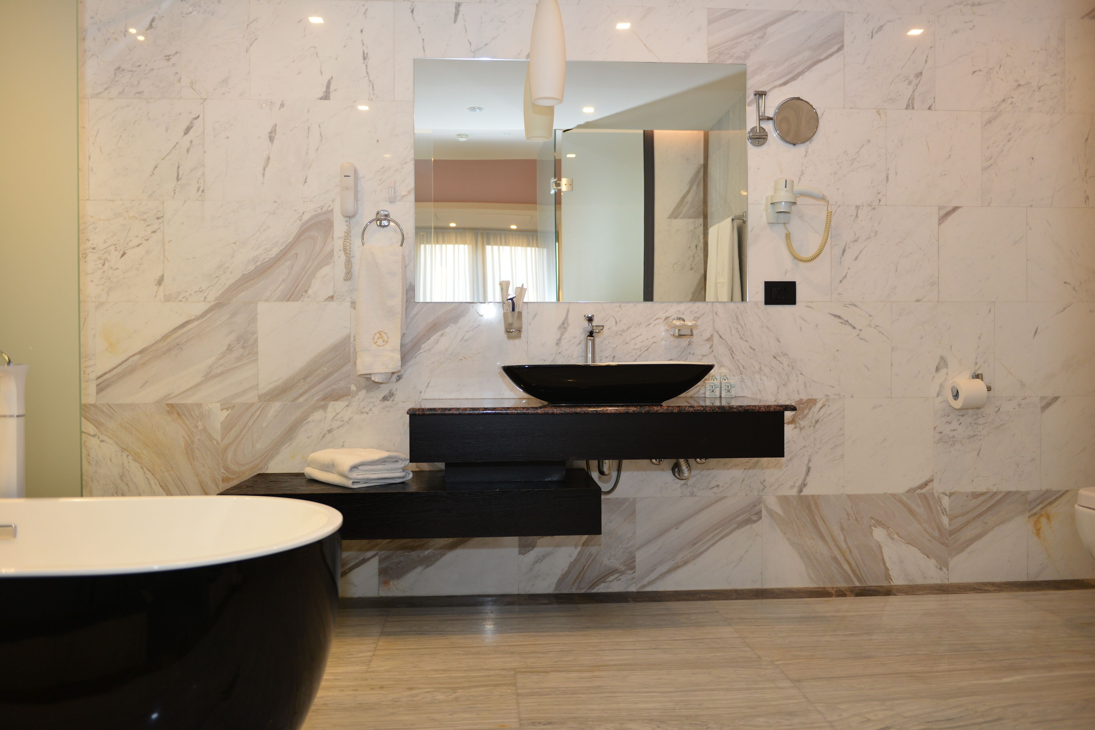 A modern bathroom with a marble wall, a black sink, a large mirror, and a black bathtub.