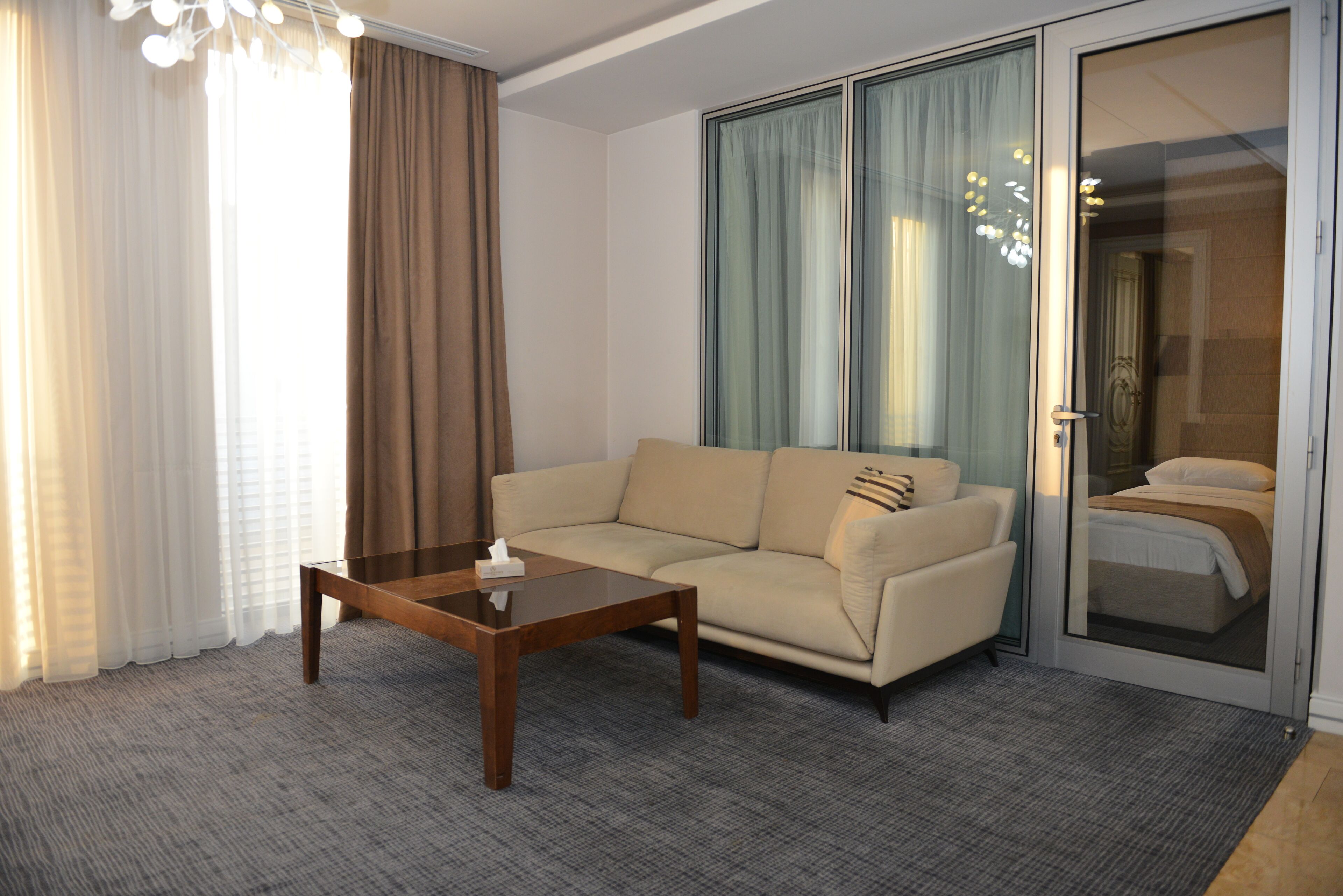 A hotel room with a sofa, a coffee table, a sliding glass door leading to a balcony, and a large window with curtains.