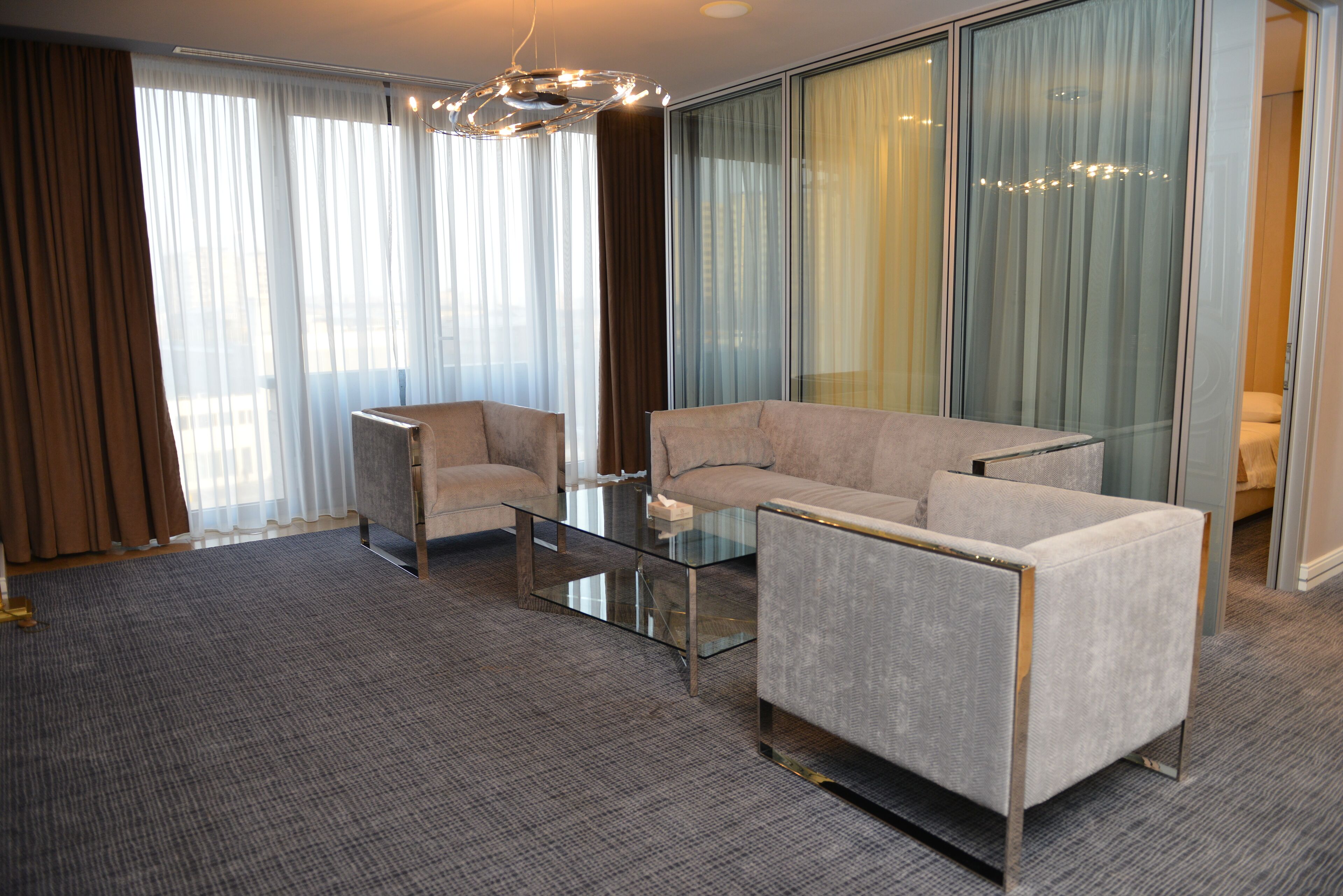 A modern hotel room with a glass coffee table, two armchairs, a sofa, and a large window with curtains.