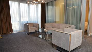 A modern hotel room with a glass coffee table, two armchairs, a sofa, and a large window with curtains.