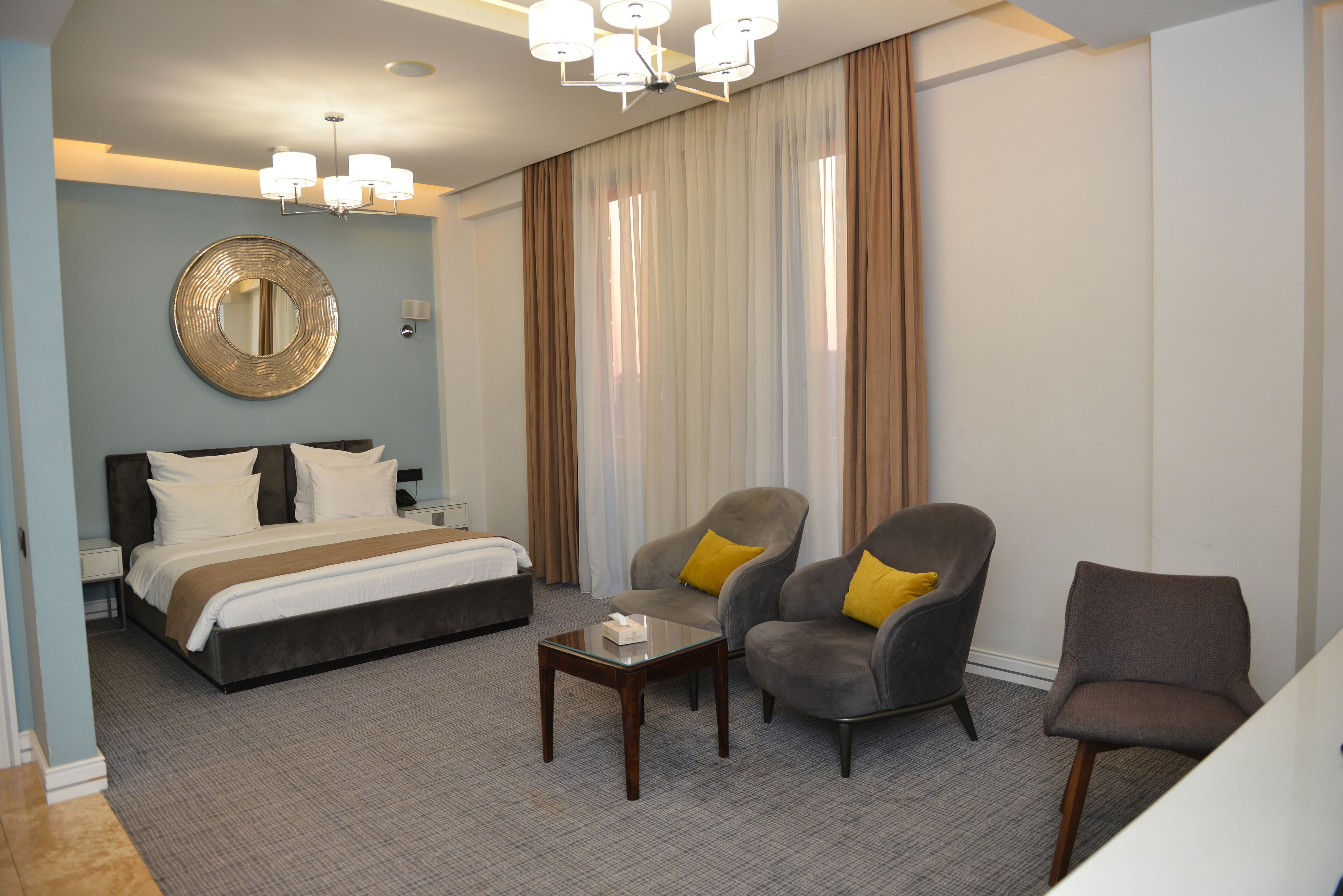 Habitación de hotel con una cama, dos sillones con cojines amarillos, una mesita y un espejo grande en la pared.