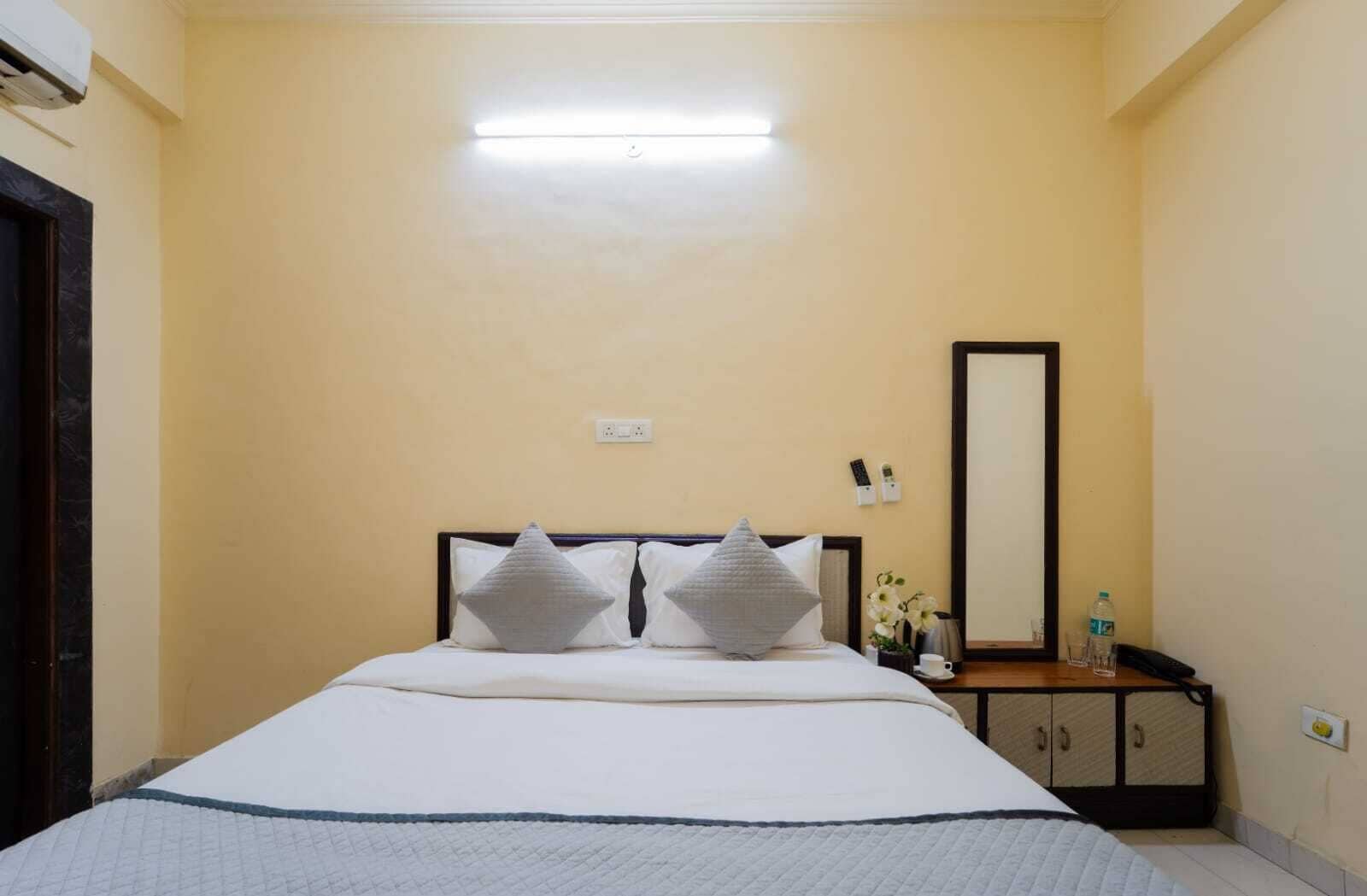 Habitación de hotel con cama, mesita de noche con lámpara, espejo y una botella de agua.