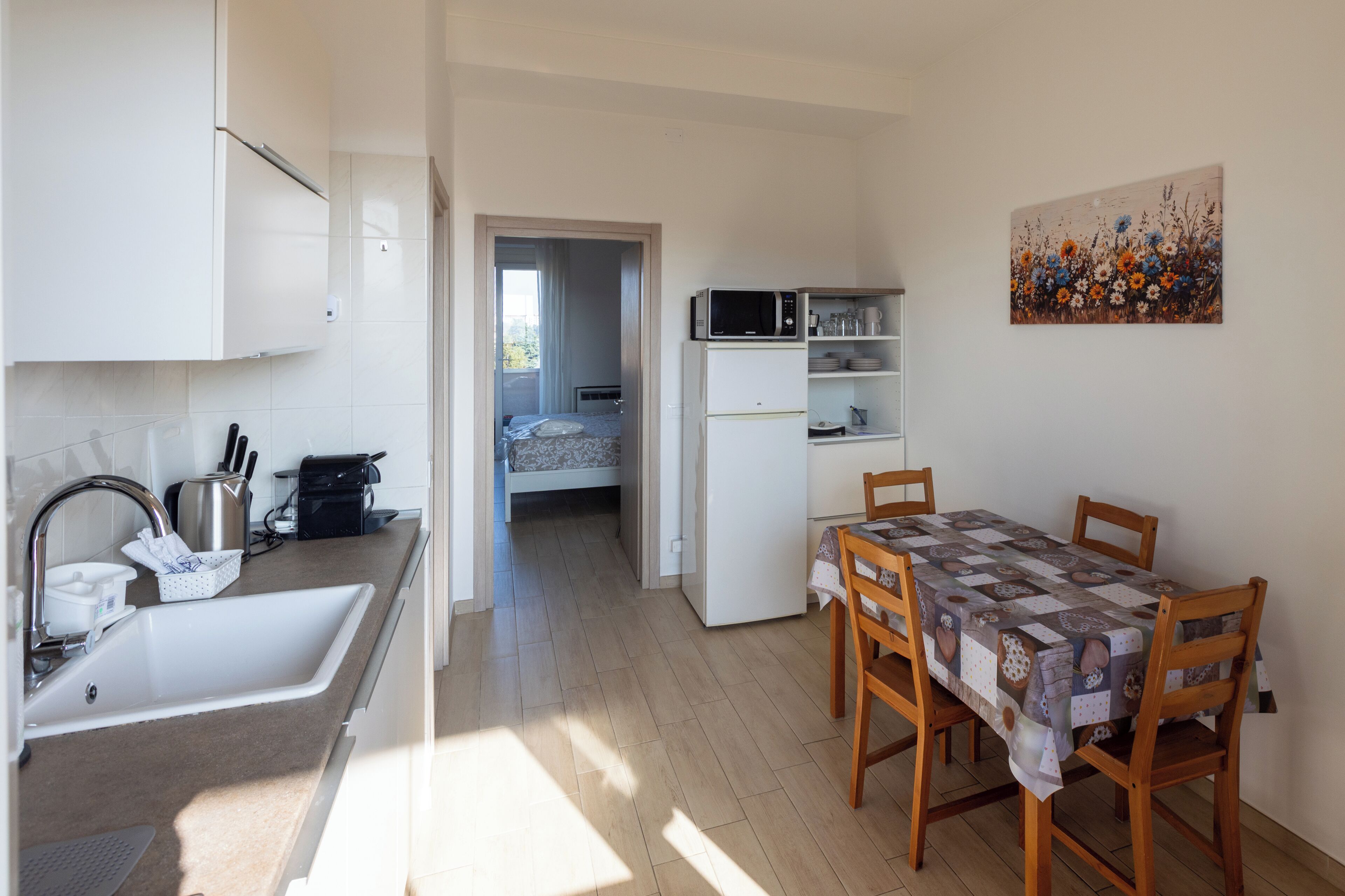 A compact kitchen with a sink, microwave, and dining table with chairs. A bed is visible in the background through an open door.