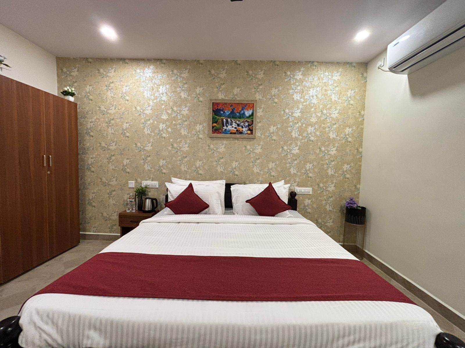 A hotel room with a large bed, a wooden wardrobe, a painting on the wall, and an air conditioning unit.