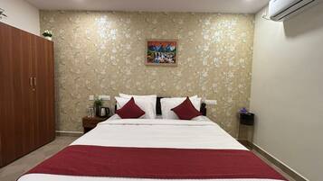 A hotel room with a large bed, a wooden wardrobe, a painting on the wall, and an air conditioning unit.