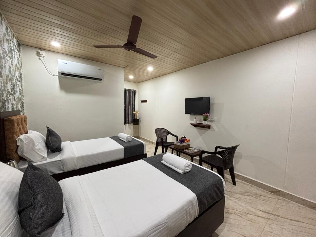 Camera d'albergo con due letti, una ventola a soffitto, un condizionatore d'aria, una TV e un tavolino con una sedia.