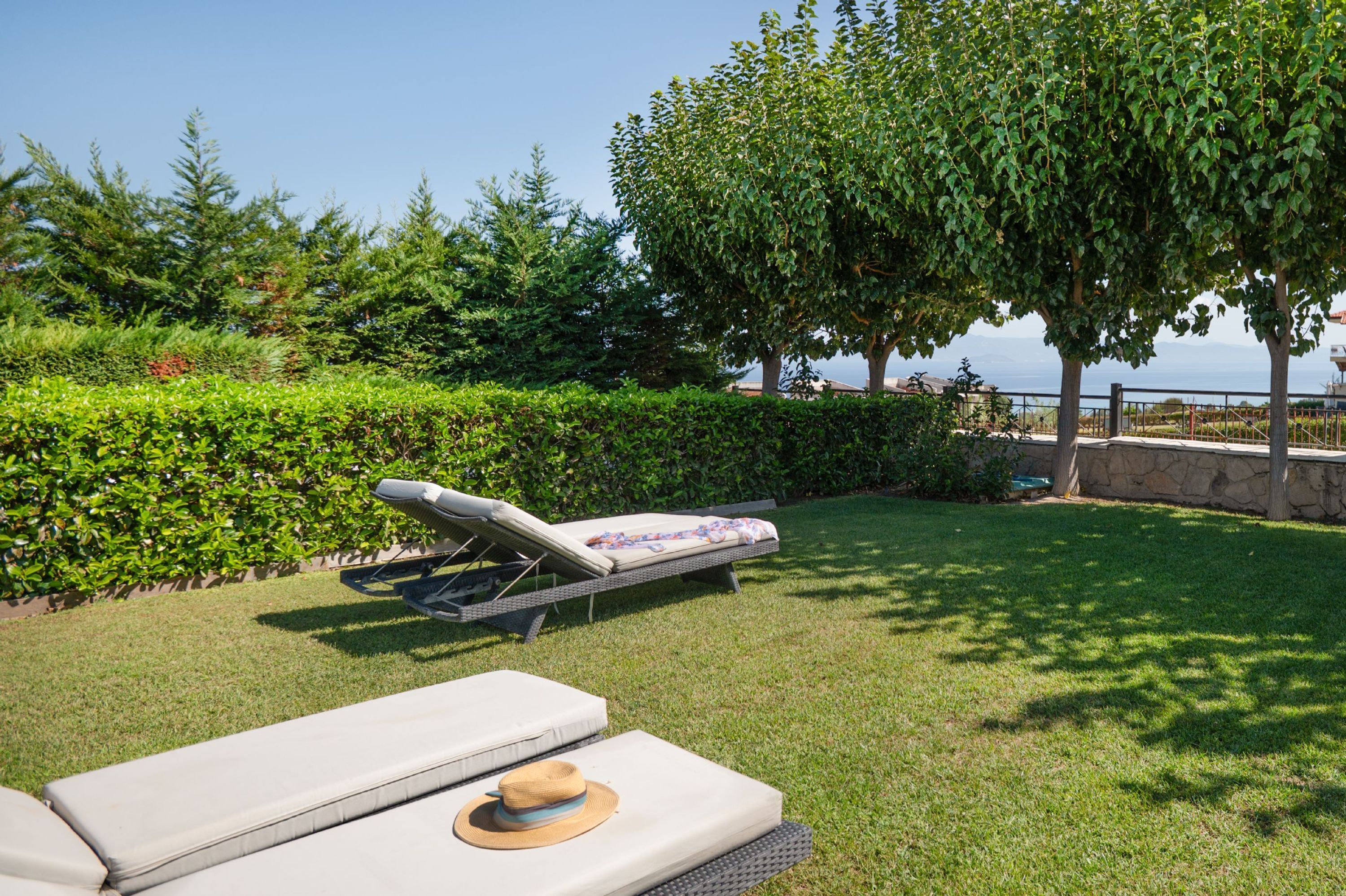 A sunlit outdoor area with a lounge chair and a small table, surrounded by well-maintained hedges and a tree.