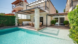 Et moderne hus i to etager med swimmingpool, udendørs møbler og en overdækket patio.