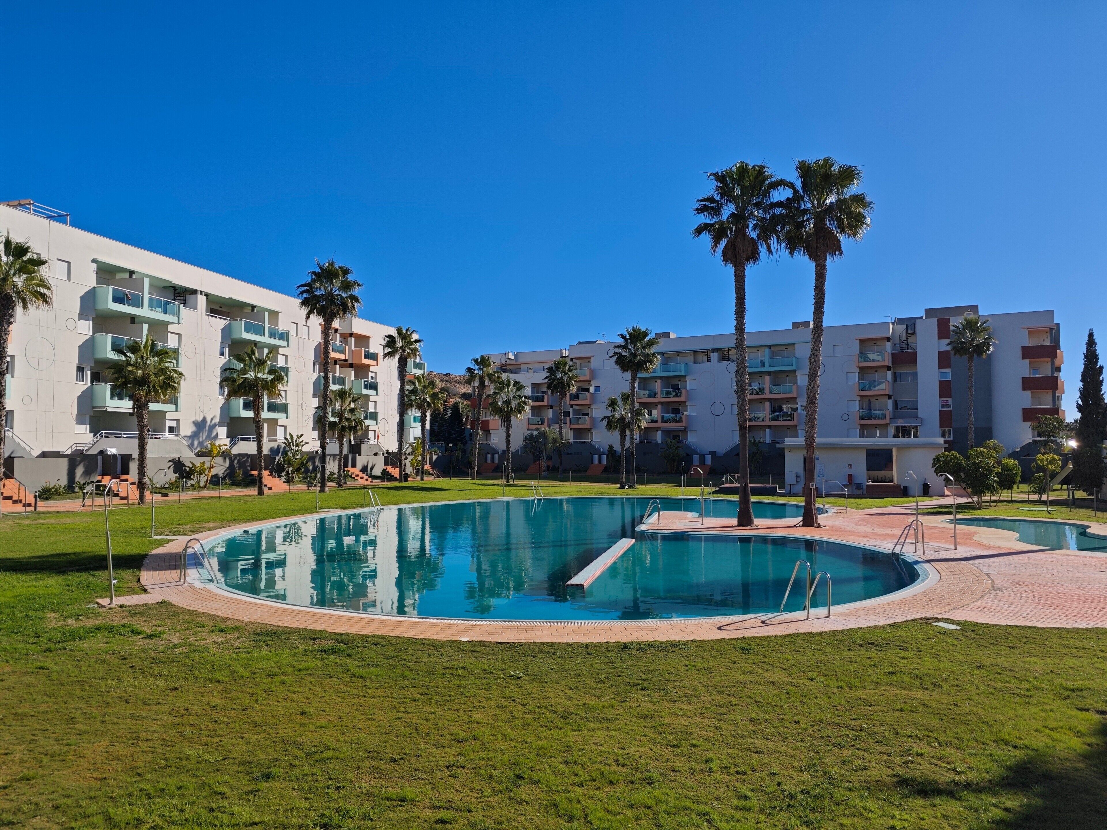Zona de piscina amb una piscina circular, palmeres i un edifici de diversos pisos amb balcons.