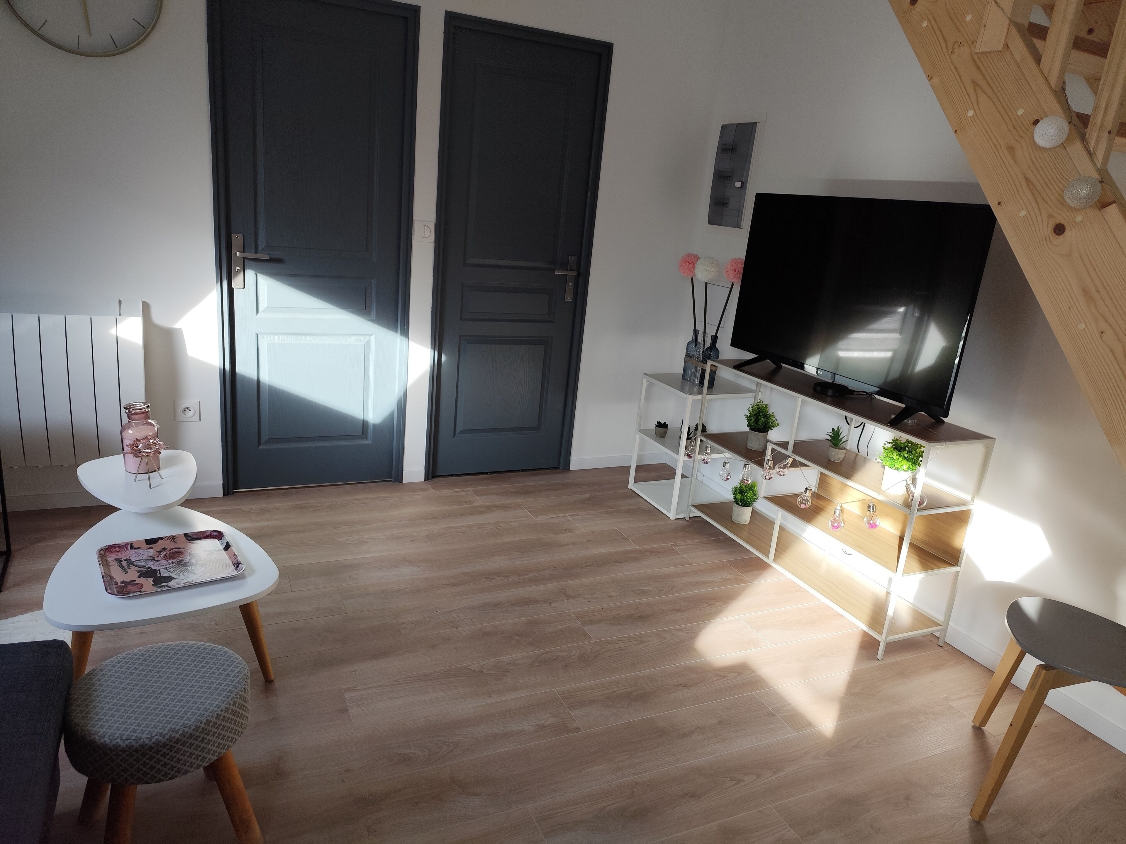 Una sala moderna con un televisor de pantalla plana sobre un mueble de madera, una mesa de centro blanca con una revista, un taburete acolchado gris y una estantería blanca con adornos.