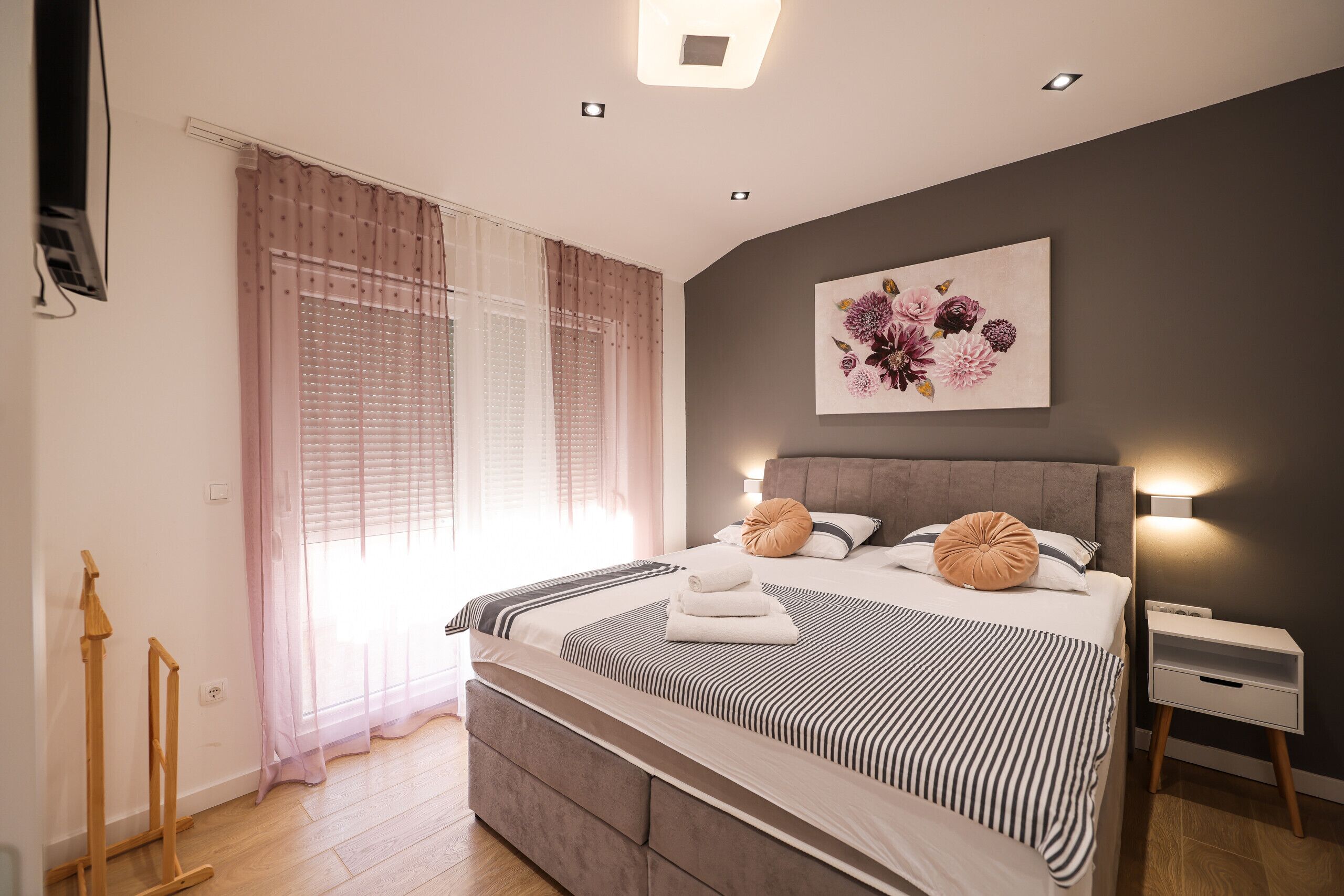 A modern bedroom with a large bed, a bedside table, a wall-mounted TV, and a window with sheer curtains.
