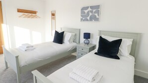A hotel room with two beds, each with white linens and dark pillows, a bedside table with a lamp, and a wall-mounted artwork. - Glynllifon 4 (Marianglas)