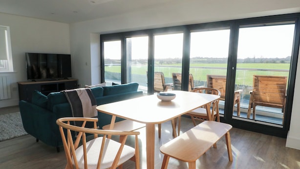 A modern living room with a large window, a sofa, a dining table, and wooden chairs. - Glynllifon 4 (Marianglas)