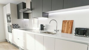 A modern kitchen with white cabinetry, a stainless steel sink, a kettle, a toaster, and a wooden cutting board. - Glynllifon 4 (Marianglas)