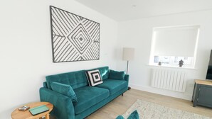 A teal sofa with a geometric-patterned pillow, a wooden side table, a floor lamp, and a view of the outside through a window. - Glynllifon 4 (Marianglas)