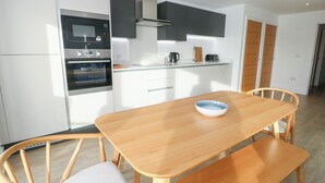 A modern kitchen with white cabinetry, a built-in oven, and a dining area with a wooden table and chairs. - Glynllifon 4 (Marianglas)