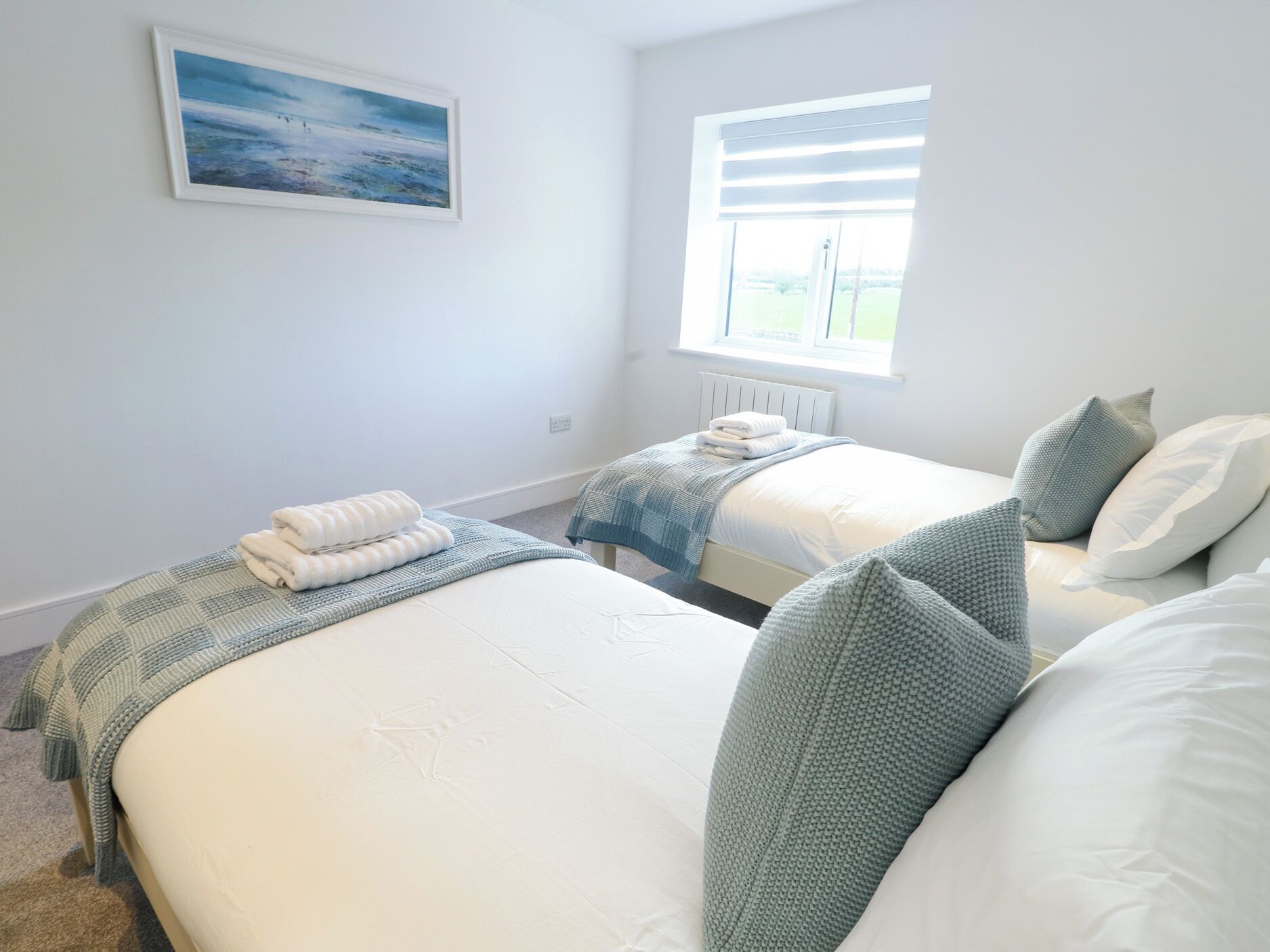 A bedroom with two beds, a window with blinds, a radiator, and a painting on the wall.