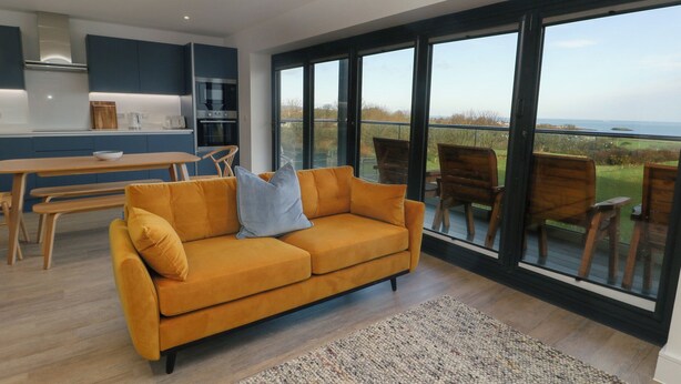 A modern living room with a sofa, a dining table, and chairs. There is a large window with a view of the outdoors. - Glynllifon 6 (Marianglas)