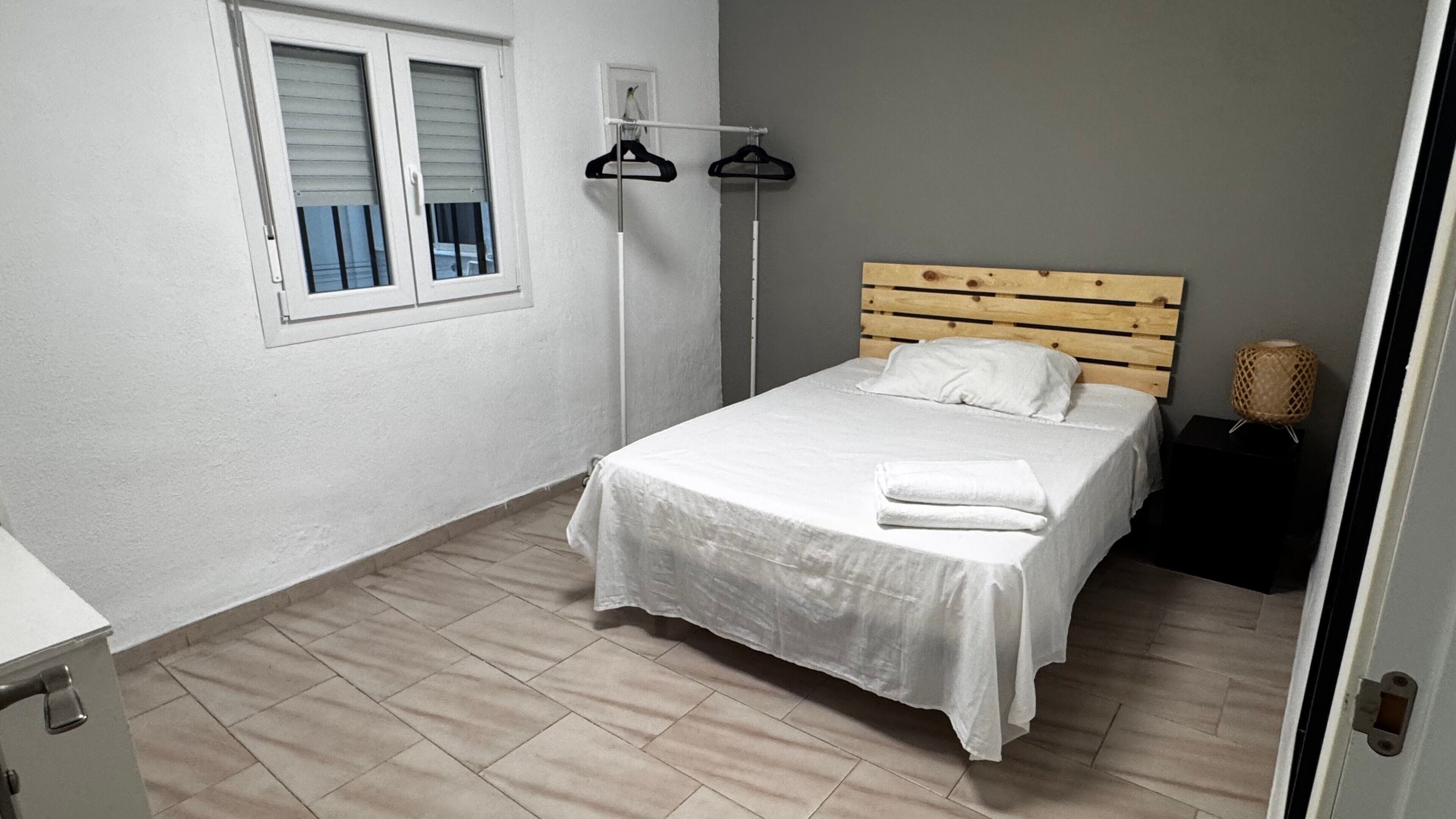A bedroom with a bed, a wooden headboard, a white wall, a window with blinds, and a floor lamp.
