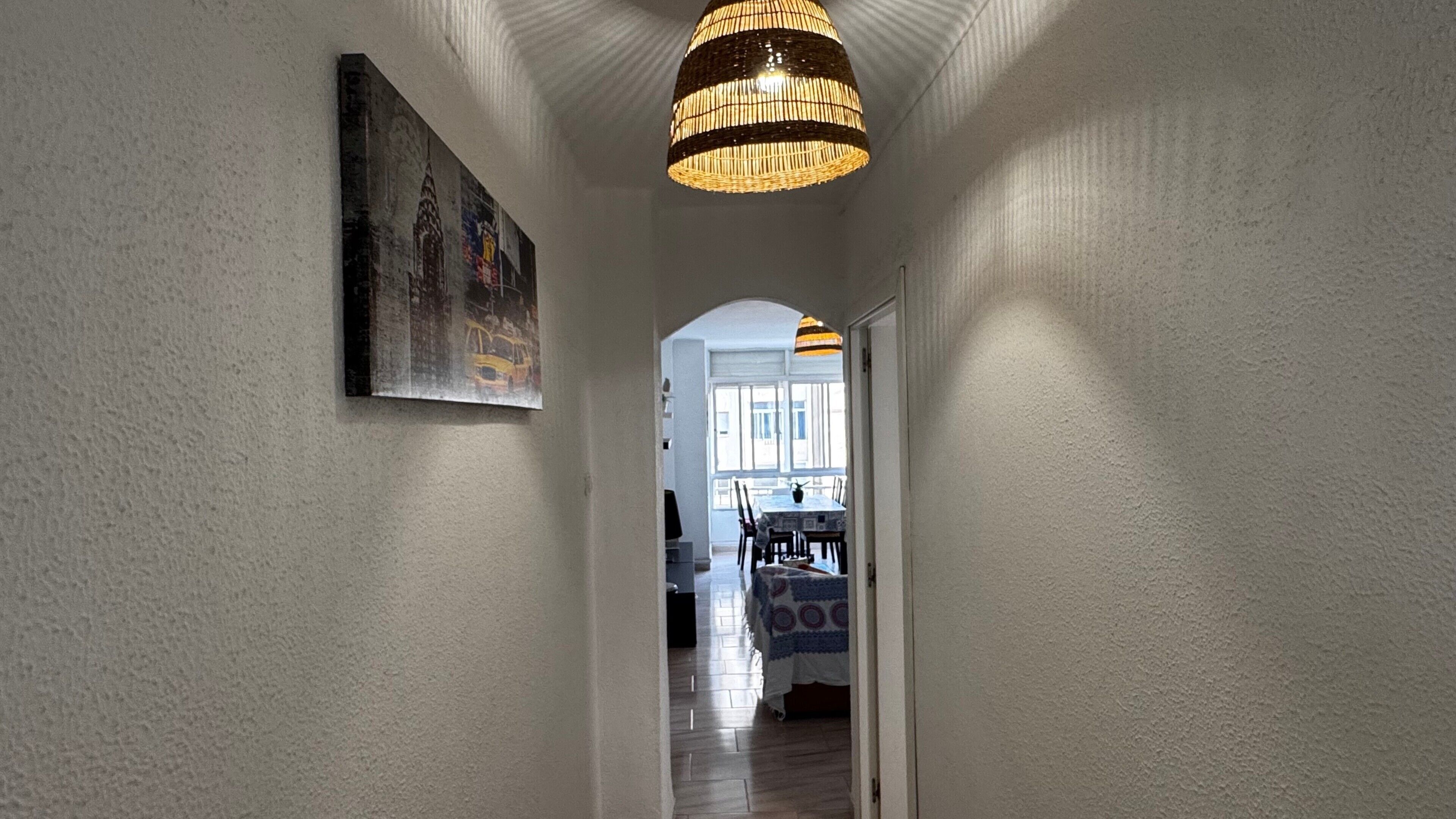 A hallway with a hanging light fixture, a painting on the wall, and a view into a dining area with a table and chairs.