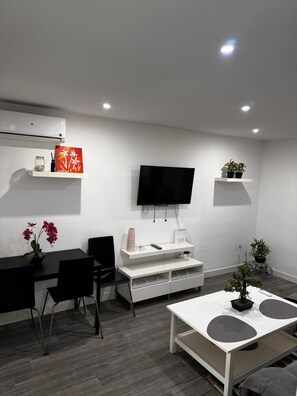 Ein modernes Wohnzimmer mit Flachbildfernseher, einem Esstisch mit Stühlen, einem Couchtisch und Topfpflanzen.