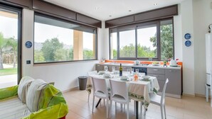 Een moderne keuken met grote ramen, een eethoek met een tafel gedekt voor vier personen, en een bed met een groen en wit beddegoed.