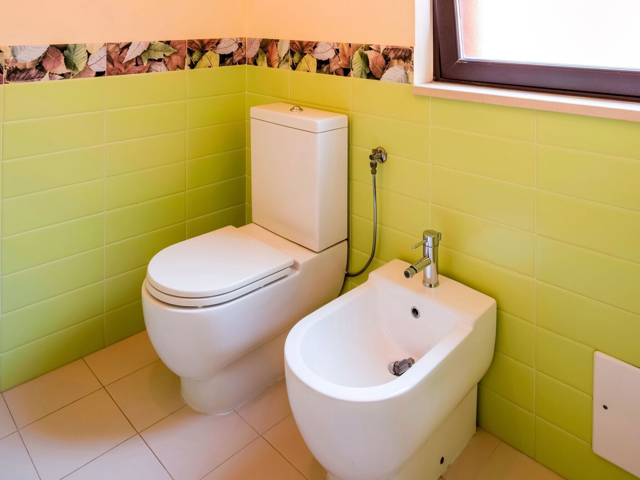 A bathroom with a toilet and bidet, green tiled walls, and a window.