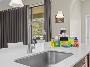 A modern kitchen with a stainless steel sink, a grey faucet, and a white countertop. There are various cleaning products on the counter, including a bottle of Dawn dish soap and a bottle of Tide detergent.