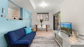 Ein modernes Wohnzimmer mit einem blauen Sofa, einem Esstisch mit Stühlen, einem Fernseher und einem großen Fenster mit Blick.