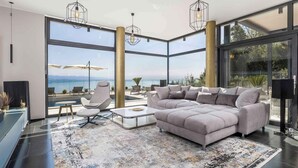 Un salon moderne avec un grand canapé d’angle, une table basse et un tapis à motifs. De grandes fenêtres offrent une vue sur la mer.