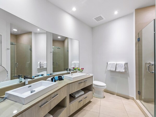 A modern bathroom with a large mirror, two sinks, a toilet, and a glass shower enclosure.