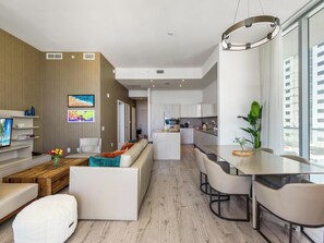A modern living room with a sofa, coffee table, and a TV. There is a kitchen area with a dining table and chairs.