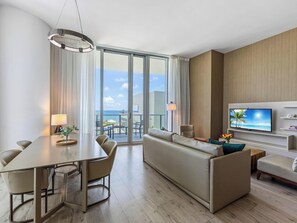 A modern hotel room with a dining area, a sofa, a TV, and a balcony with a view of the ocean.