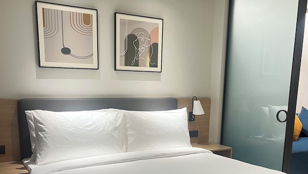 Habitación de hotel moderna con una cama grande, dos cuadros enmarcados en la pared, una mesita de noche con una lámpara y una puerta corredera de vidrio a la derecha.