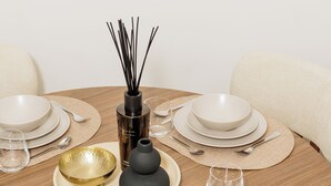 Een ronde houten eettafel, gedekt met witte borden, kommen en bestek, met een gouden schaal en een zwarte vaas met rietstelen.