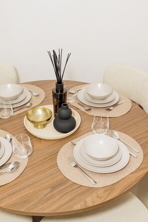 Una mesa de madera redonda dispuesta con platos, fuentes y cubiertos blancos, junto a un cuenco dorado y un florero negro con ramas de caña.