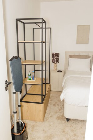 A bedroom with a bed, a wooden nightstand, a wall-mounted shelf, a vacuum cleaner, and a door. - Stylish & modern studio at Sacré-Cœur 1, Dakar (Dakar)