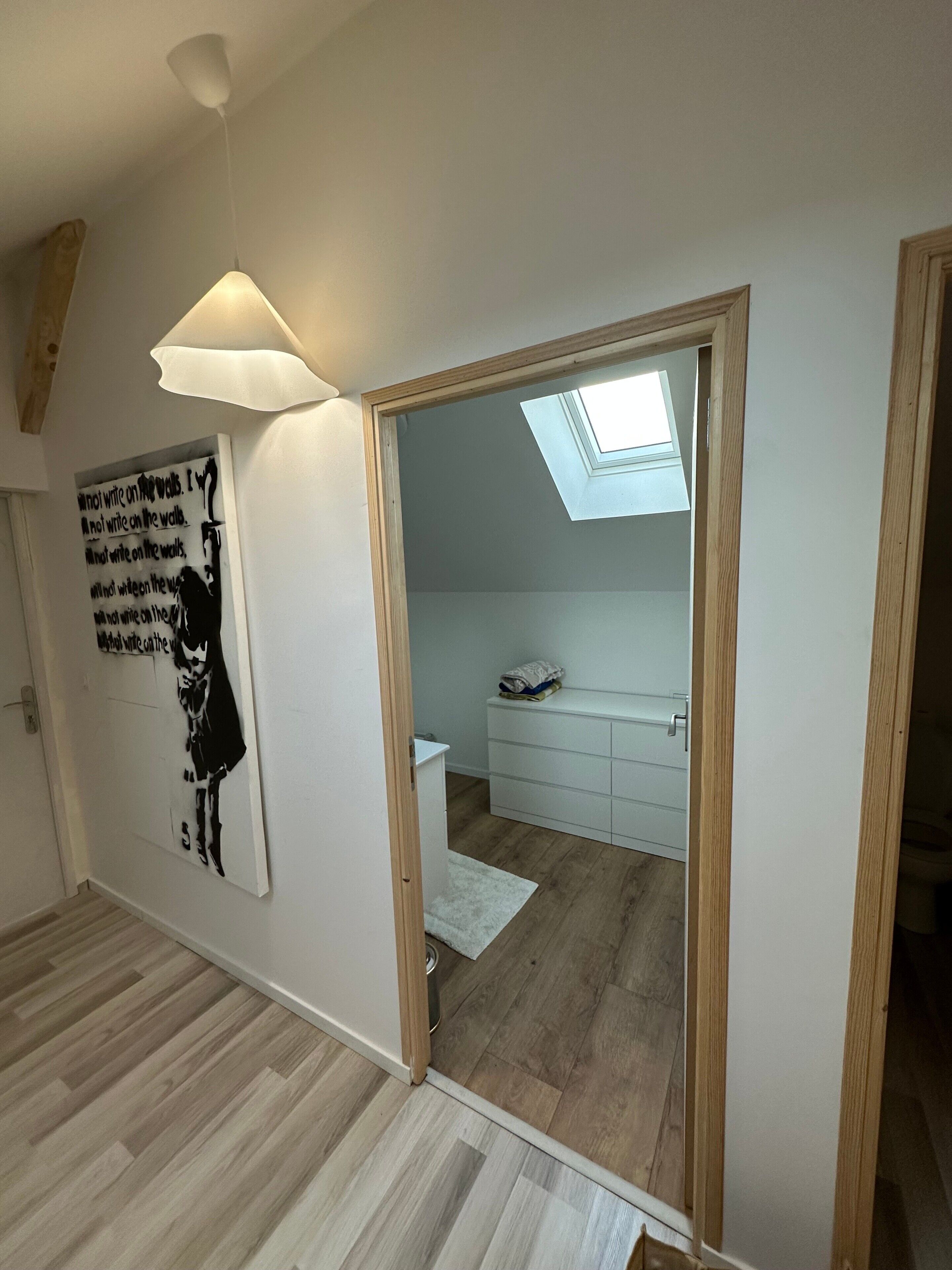 A room with a skylight, a white wall with a black and white poster, and a wooden floor.