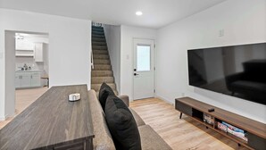A modern living room with a large flat-screen TV, a wooden dining table, and a staircase leading to a kitchen. - Rivers Edge Retreat For 10ppl (Dayton)