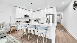 A modern kitchen with white cabinetry, a central island with bar stools, and a stainless steel refrigerator. - The Grove Holly 133- 1 Bedroom - Lazy River - Seascape Resort (Miramar Beach)